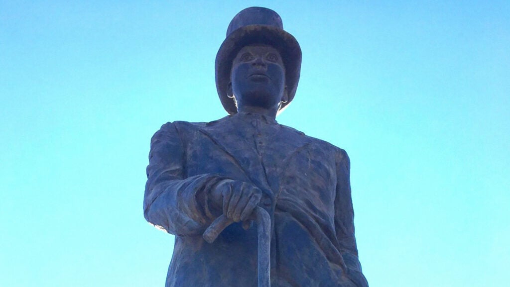 Close up of the upper half of a statue, a man named Pixley ka Seme. He is wearing a top hat and a jacket with a button-up shirt; he is holding a cane in his right hand. The statue is metal (like bronze) and has a dark brownish/black color.