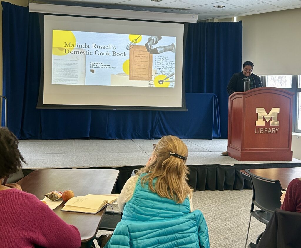 Jessica Kenyatta Walker stands at a podium on a stage. Behind her a slide shows the cover of Malinda Russell's "A Domestic Cookbook"