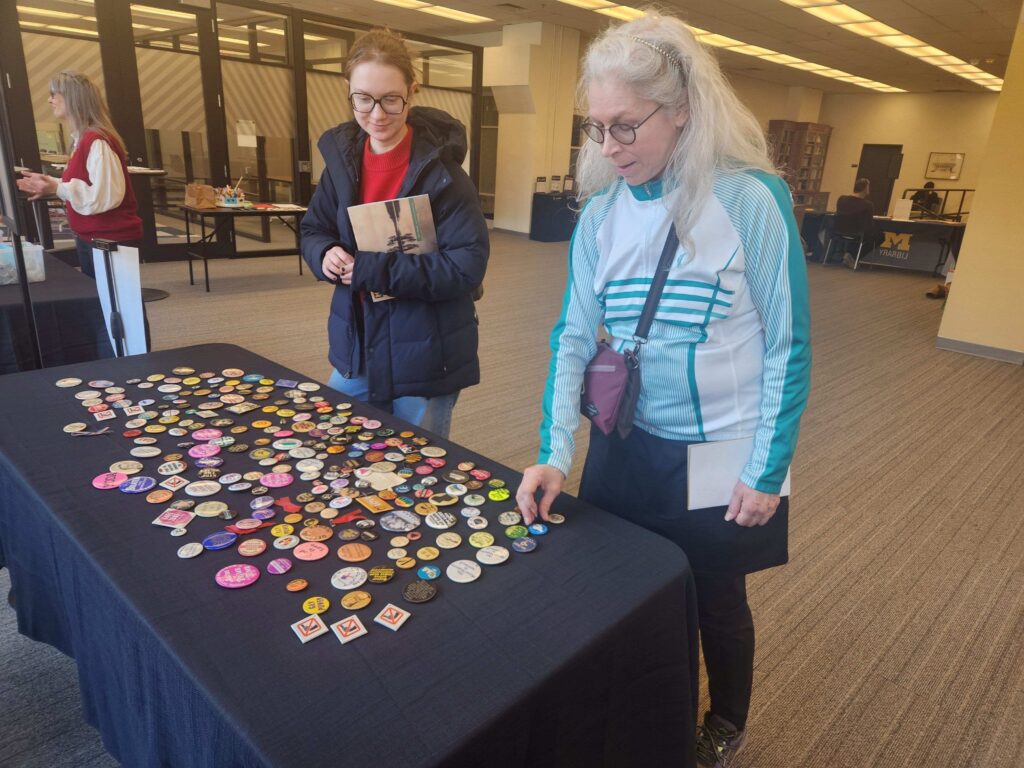 Two people look at an array of activist pinback buttons on a table. The buttons have slogans such as "Gay Power," God is Love," "Stop the Draft", and "No Justice No Peace."