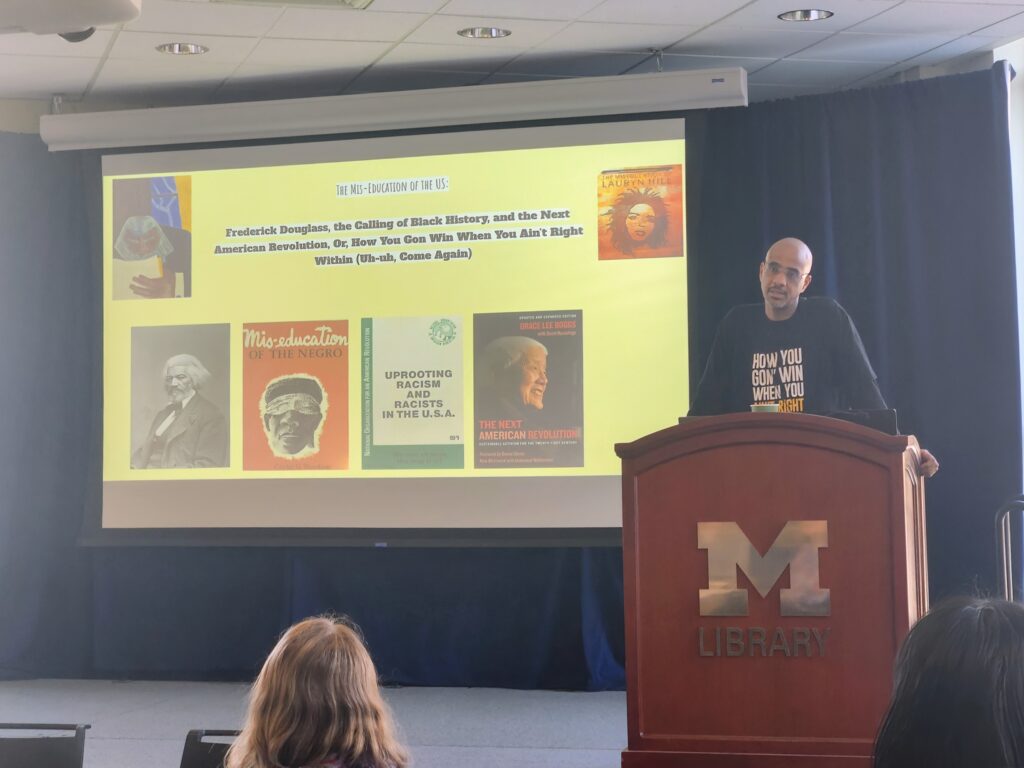 Stephen Ward stands at a podium in front of a projection of a slide that reads "The Mis-Education of the US: Frederick Douglass, the Calling of Black History, and the Next American Revolution, Or, How You Gon Win When You Ain't Right Within (Uh-uh, Come Again)." Also visible on the slides are: a photo of Frederick Douglass, a painted Portrait of Frederick Douglass, Book covers of "Mis-education of the Negro," "Uprooting Racism and Racists in the U.S.A.," and "The Next American Revolution," and the album cover of "The Miseducation of Lauryn Hill."